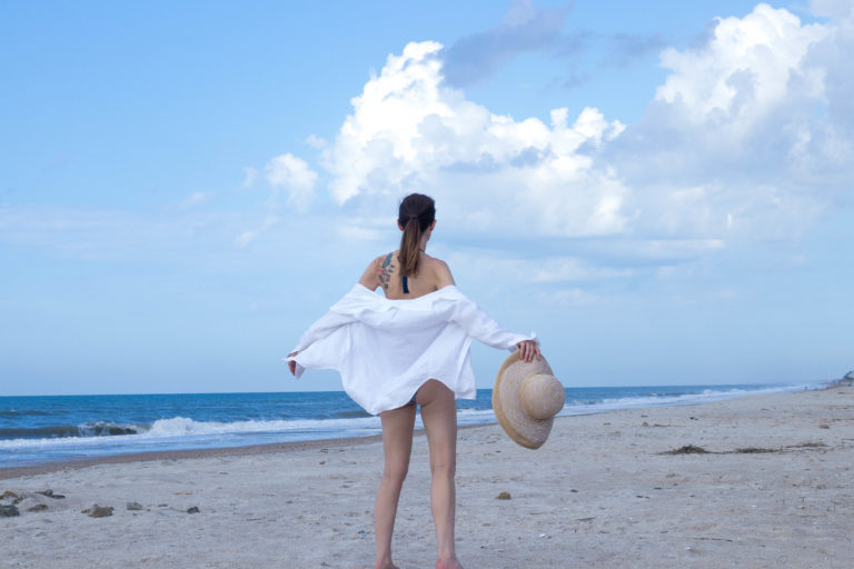 Michelle is standing on Topsail Beach North Carolina wearing a bikini covered with an open white linen shirt, her arms outstretched facing the bright blue sky and bursting white fluffy cloud. She wants to protect her Mother Earth.